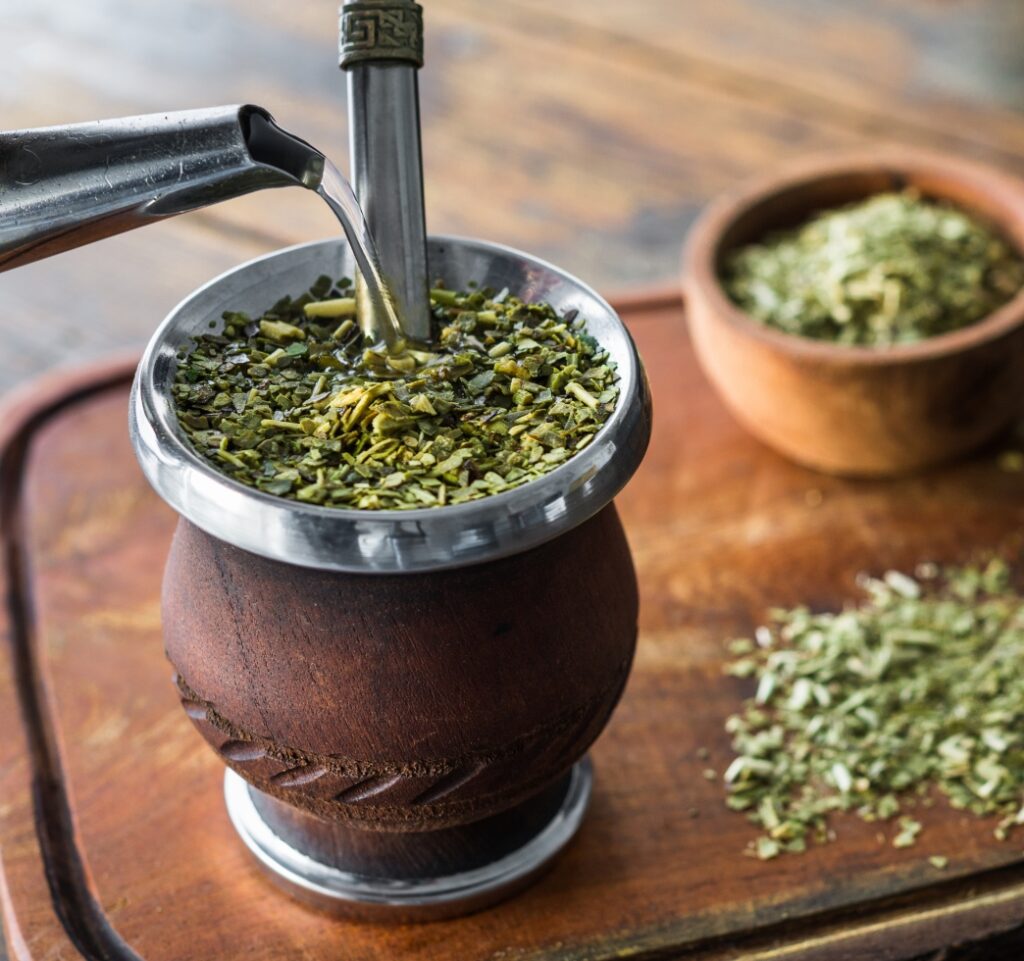 This image shows a traditional yerba mate cup made from a gourd, filled with green mate leaves and fitted with a metal bombilla for sipping. To the side, a wooden bowl holds extra dried leaves, all set on a rustic wooden table.